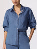 Classy denim outfit close-up showing a medium-wash button-front shirt with a spread collar, chest pocket, and rolled sleeves over a dark belt.
