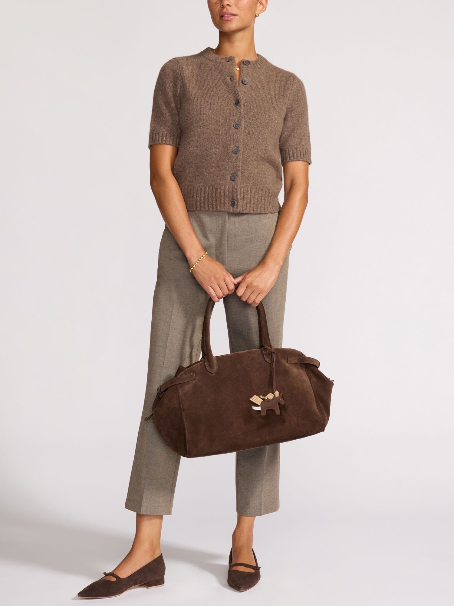 Woman holding a large dark brown suede tote bag with a horse charm, wearing a taupe cropped button-front knit cardigan and olive trousers.