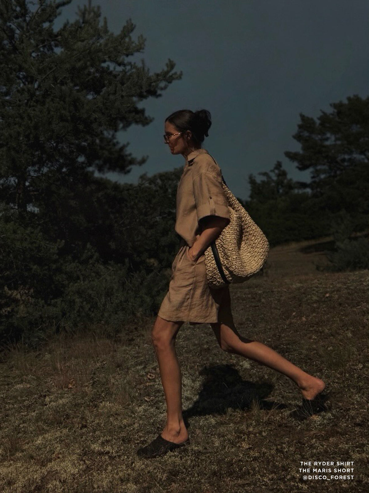 Woman walking through a wooded landscape at dusk wearing a beige linen shirt dress and carrying a woven straw tote bag