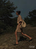 Woman walking through a wooded landscape at dusk wearing a beige linen shirt dress and carrying a woven straw tote bag
