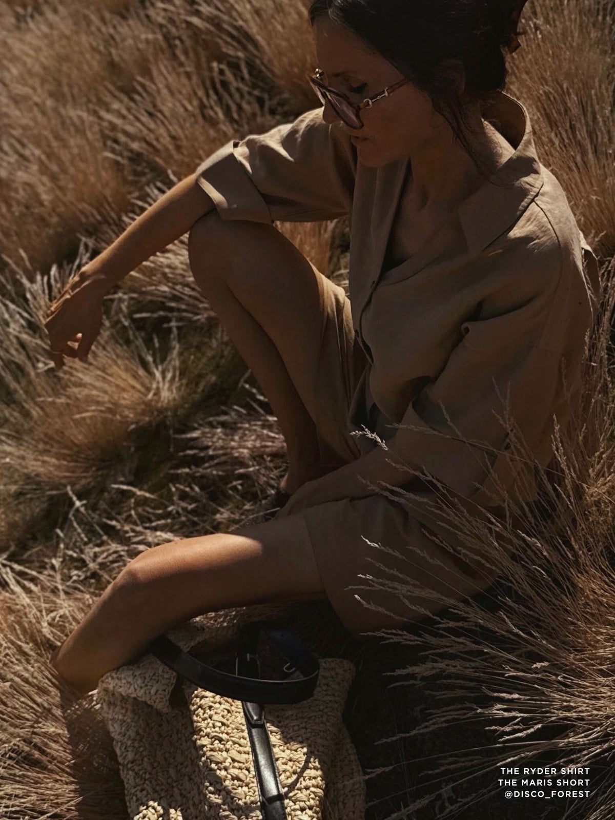 Woman crouching in tall dry grass wearing a beige linen shirt and matching shorts in golden-hour light