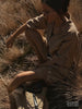Woman crouching in tall dry grass wearing a beige linen shirt and matching shorts in golden-hour light