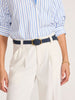 Close-up of a navy leather belt with a gold buckle worn over white pleated trousers and a blue striped button-down shirt.