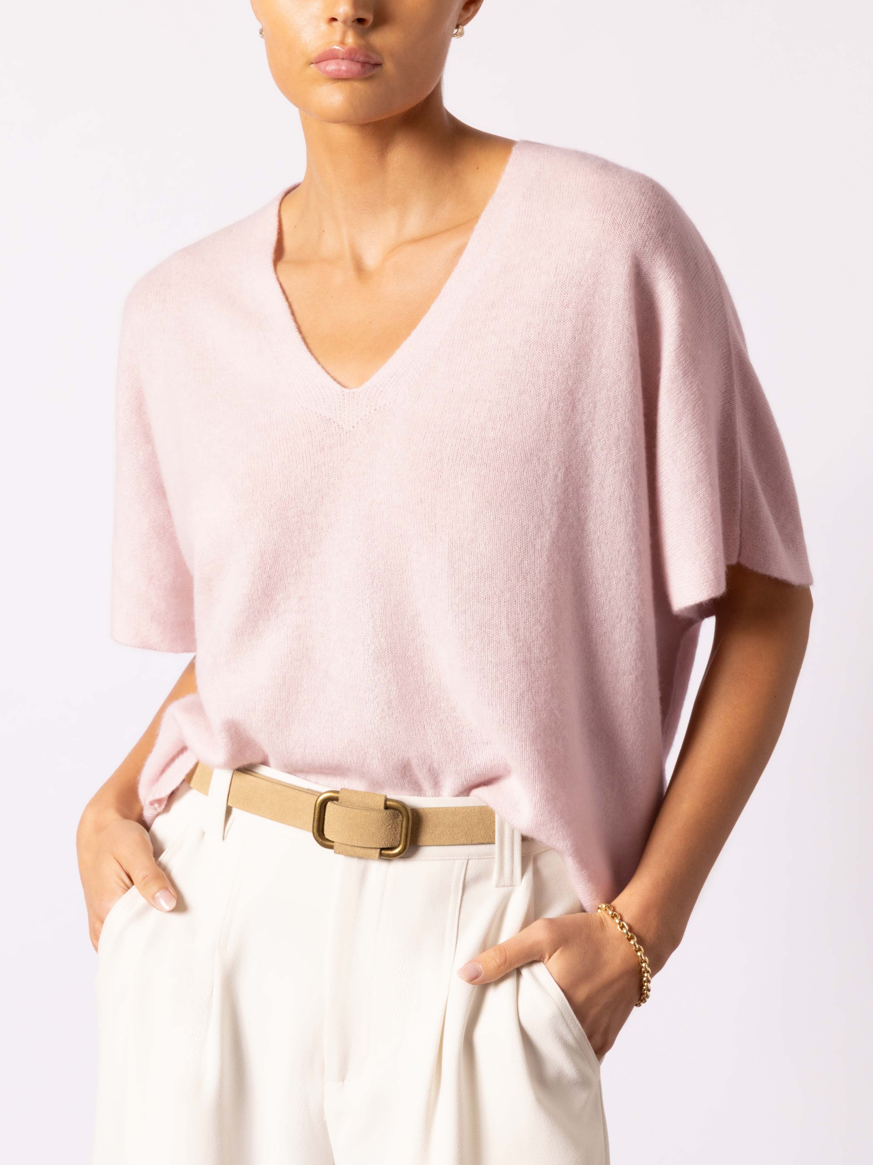 Woman wearing an elegant pink cashmere v-neck top with drop shoulders, tucked into cream wide-leg trousers with a tan belt.