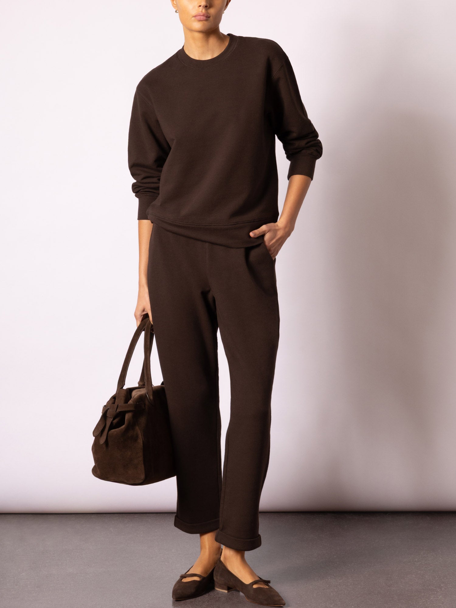 Woman wearing a matching dark chocolate brown crewneck sweatshirt and wide-leg jogger set, styled with a suede tote and mary jane flats.