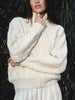 Woman wearing The Avalon Bomber Cardigan, a chunky cream cable-knit zip-up sweater, against a leafy backdrop