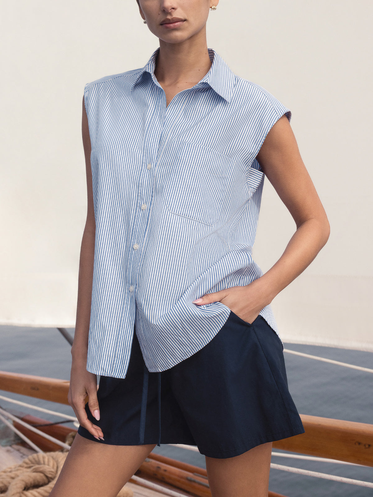 Woman wearing The Everyday Sleeveless Stripe Shirt, a blue-and-white striped button-up top, standing on a boat deck