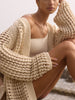 Woman wearing The Keifer Cardigan, a chunky cream open-knit cardigan, seated outdoors