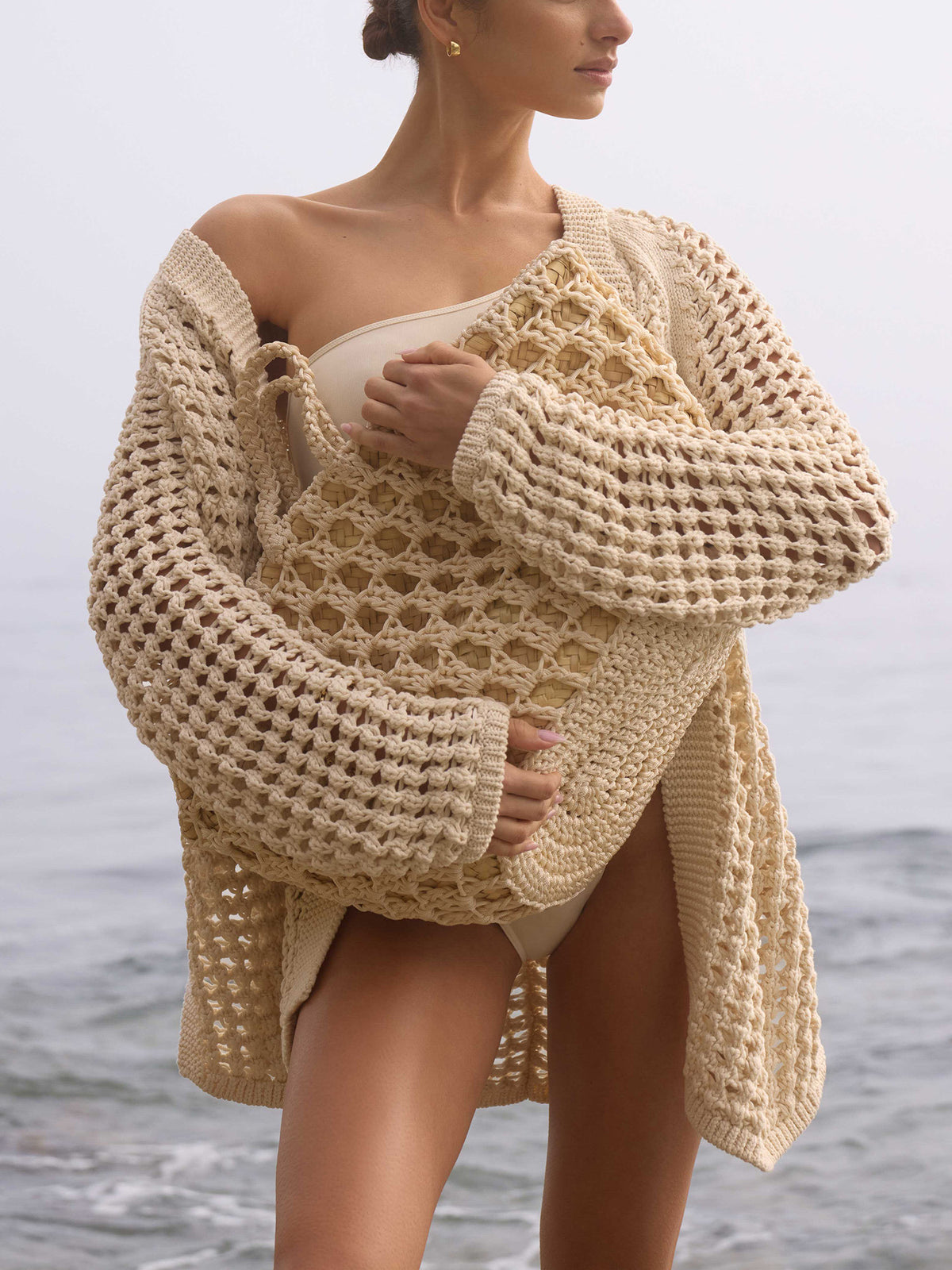 Woman on the beach wearing The Keifer Cardigan, a chunky cream crochet cardigan, over a swimsuit