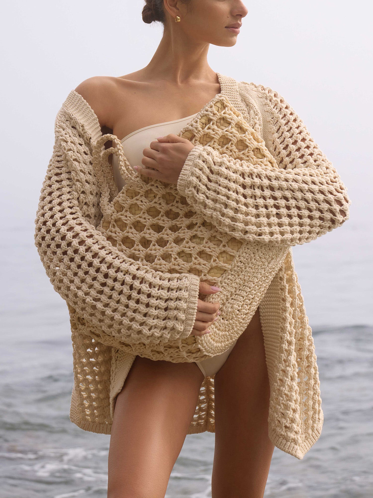 Woman on the beach wearing The Keifer Cardigan, a chunky cream crochet cardigan, over a swimsuit
