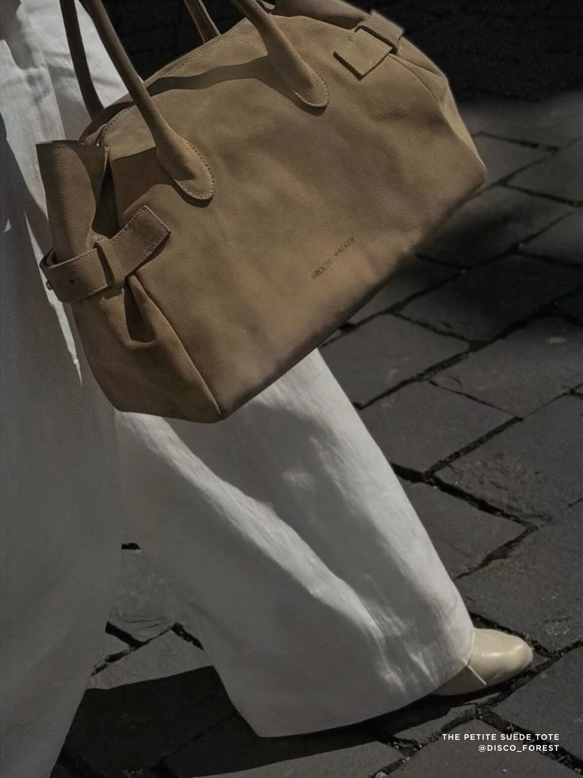 The Petite Suede Tote in tan carried at a model's side over white wide-leg trousers on a cobblestone street