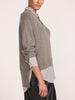 Side profile of a woman wearing a pullover v-neck sweater in otter melange with flint stripe, layered over a striped shirt with black trousers.