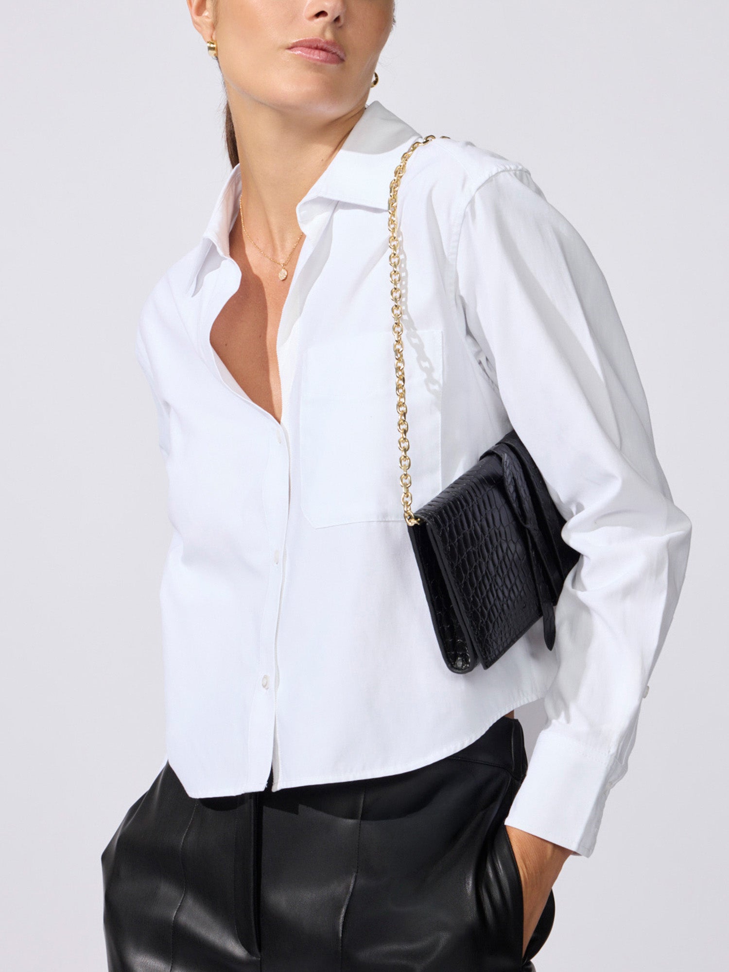 Close-up of a black crocodile-embossed clutch bag on a gold chain strap, worn over a white button-down shirt and black leather trousers.