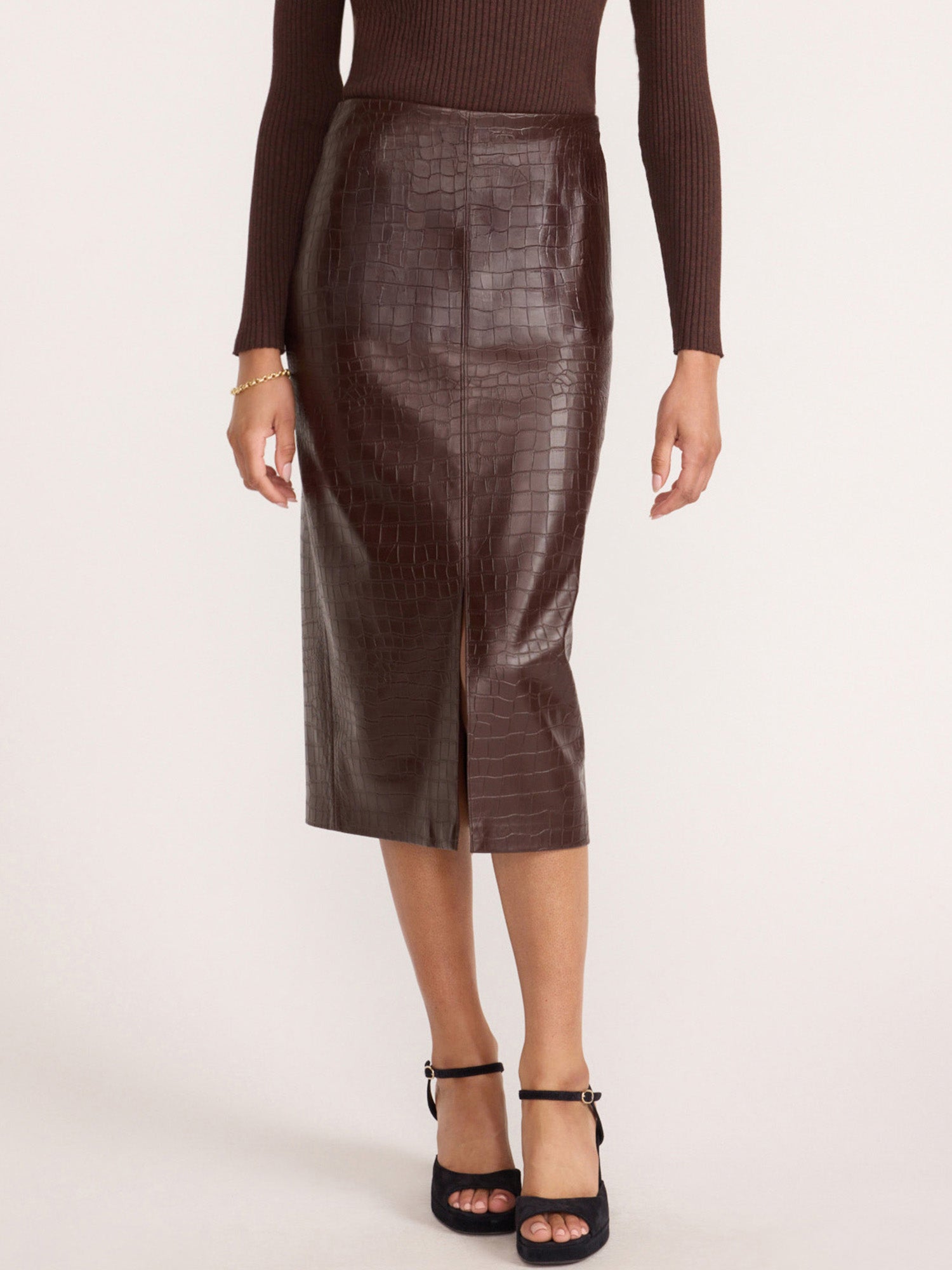 Woman wearing a dark brown crocodile-embossed midi skirt with a front slit, paired with a ribbed knit top and black heeled sandals.