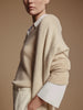 Close-up of a beige v-neck sweater layered over a white collared shirt with the white cuffs showing, worn with cream trousers