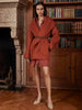 Belted wrap coat in rust orange, worn with a matching skirt and beige heels, standing in front of a bookshelf