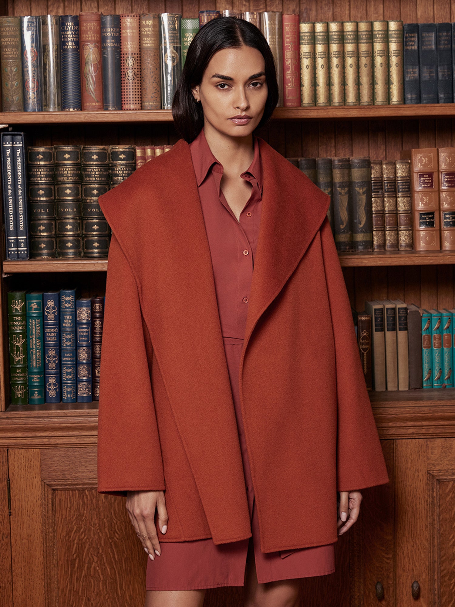 Oversized wrap coat in rust orange, worn over a matching collared shirt and shorts set, styled in front of a bookshelf