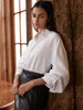Woman wearing a white long-sleeve button-up shirt with a black leather skirt, leaning against a patterned curtain.