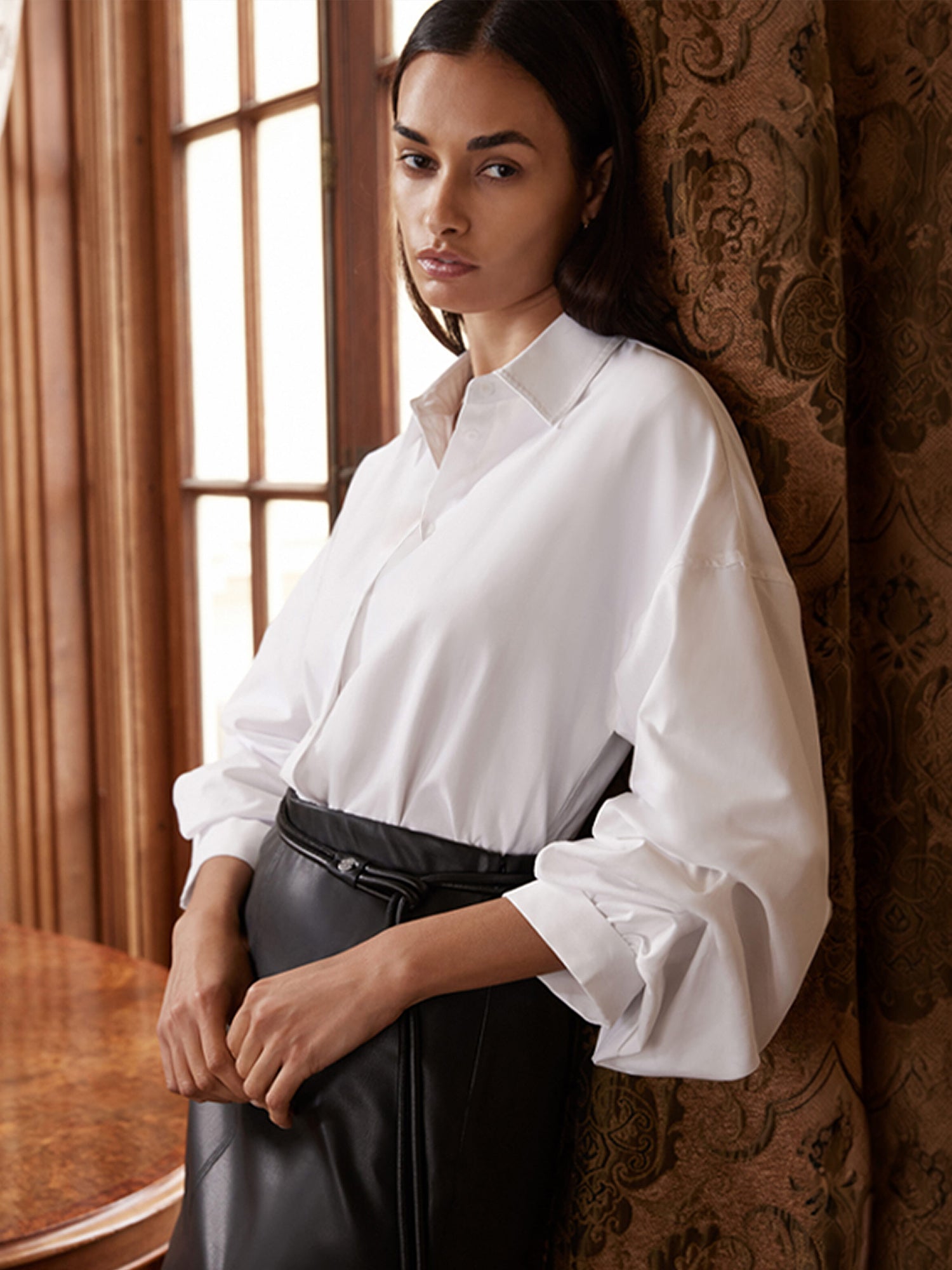 A woman wearing a white button-up blouse with voluminous sleeves and a black leather skirt, posing in an elegant room with patterned brown curtains and a window.