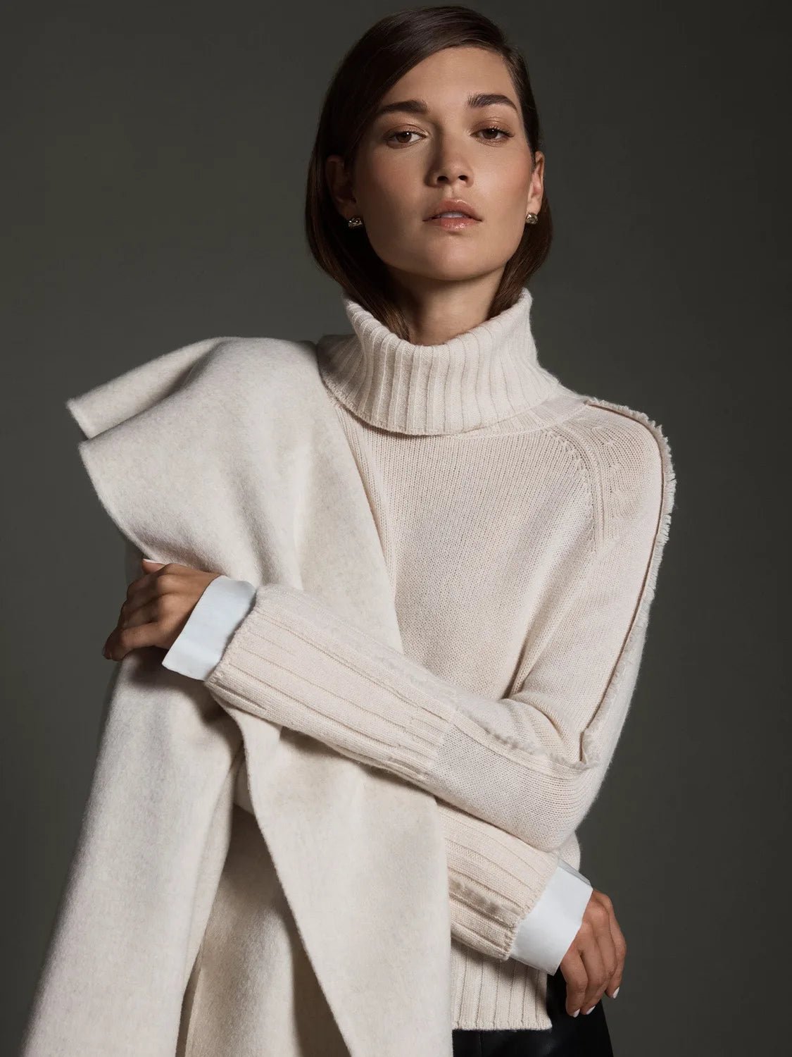 Model wearing a cream turtleneck layered sweater with white satin cuffs and a draped wool wrap.