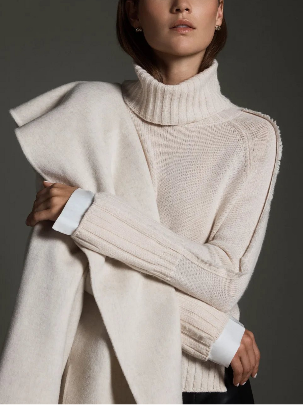 Model wearing a cream turtleneck layered sweater with white satin cuffs and a draped wool wrap.