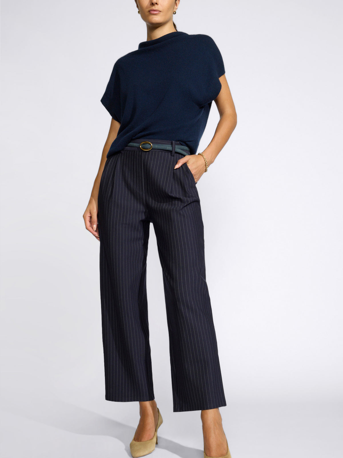 Model wearing a navy short-sleeve mock-neck top with navy pinstripe cropped trousers and beige heels, against a light gray studio background.