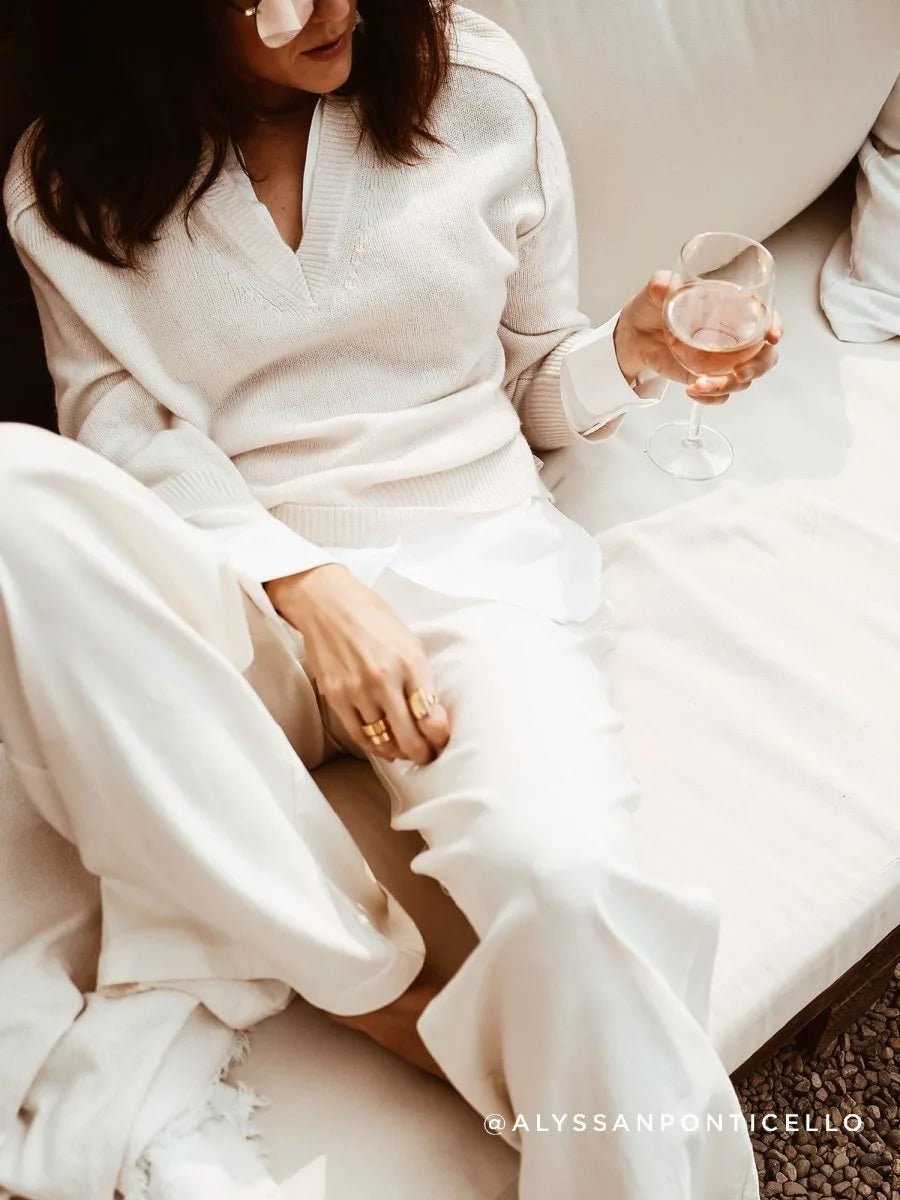 Person in a cream layered V-neck sweater and white pants lounging with a glass of rosé.
