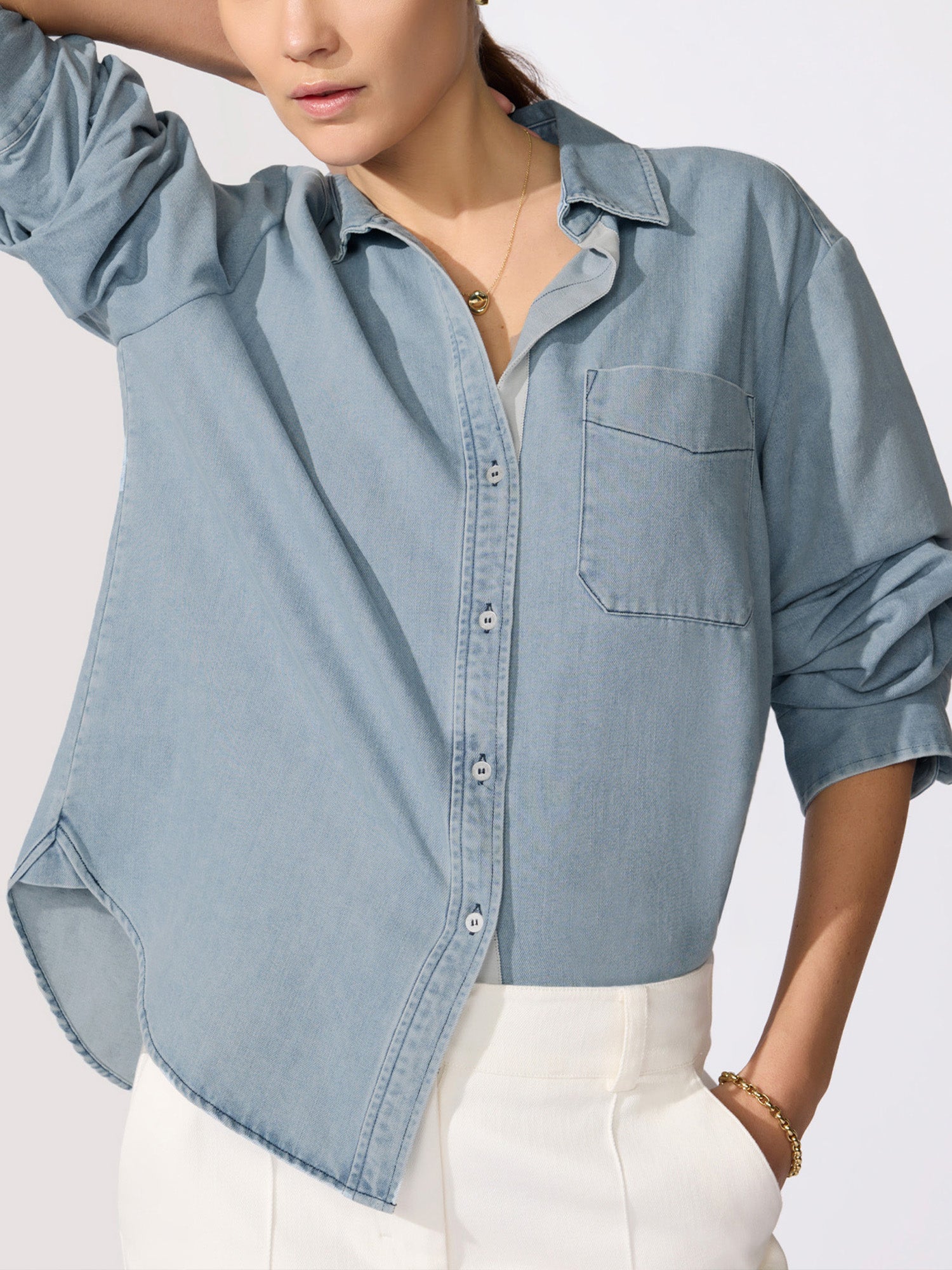 Woman wearing an open light blue denim shirt over white pants with her hand raised to her hair