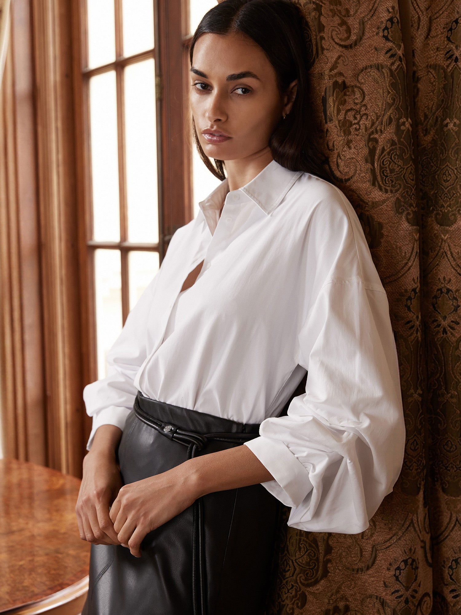 Woman wearing a white long-sleeve button-up shirt with a black leather skirt, leaning against a patterned curtain.