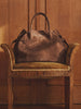 Brown suede tote bag with double top handles, displayed on a vintage wooden armchair